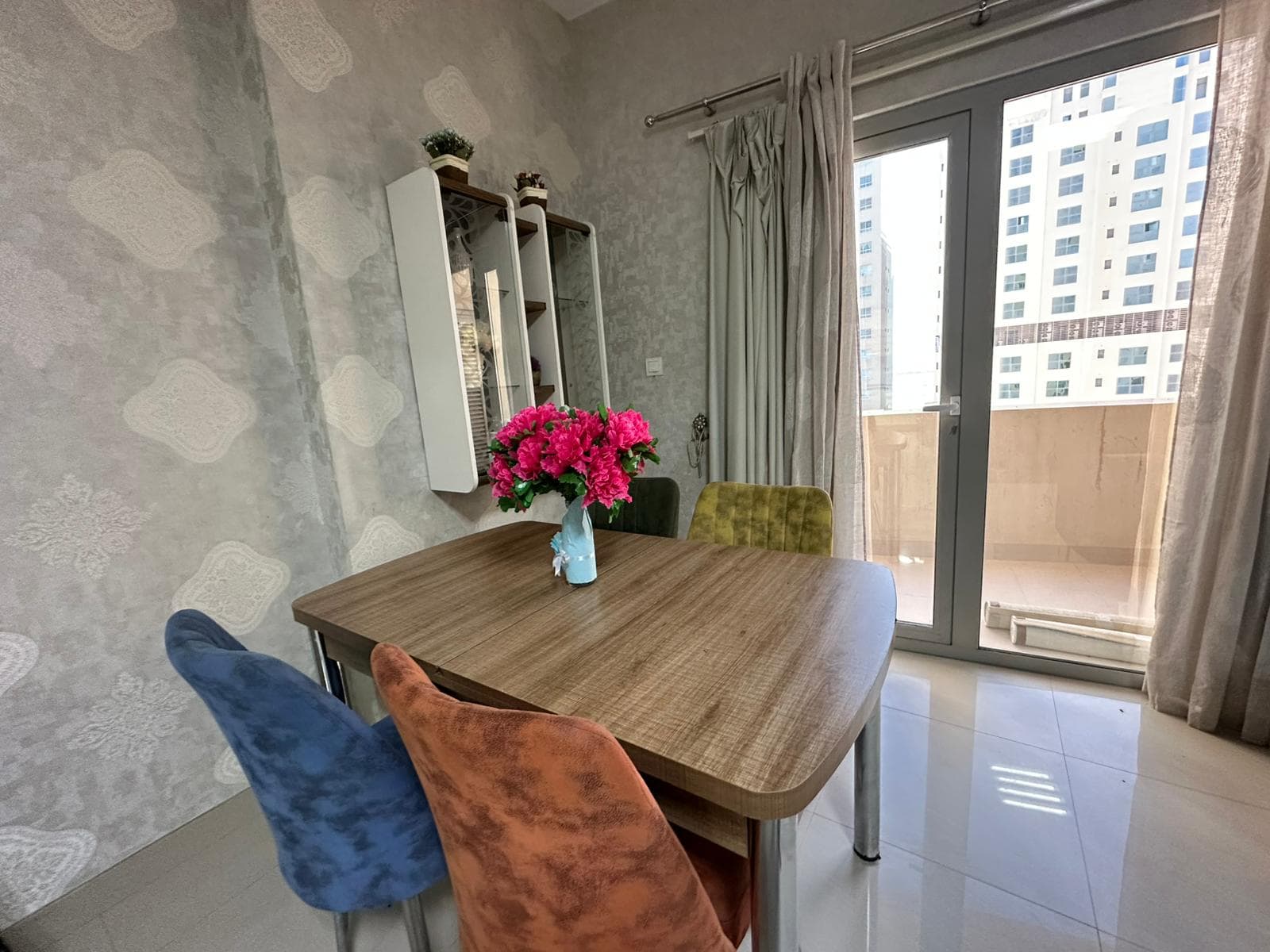 Dining area with a wooden table, a blue, orange, and green chair, and a vase of pink flowers. Background includes a balcony view and a patterned wallpaper.