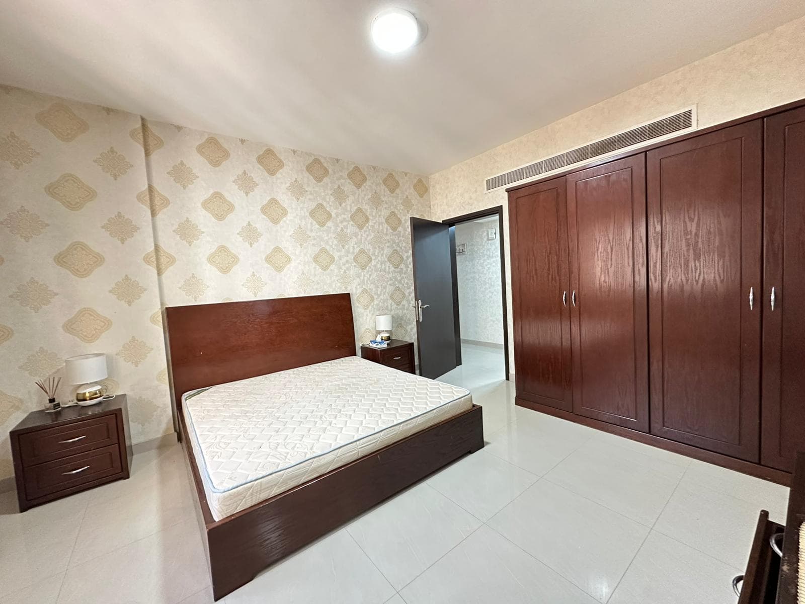 Bedroom with double bed, light patterned wallpaper, dark wooden wardrobe and side tables, white tiled floor, and overhead lighting.