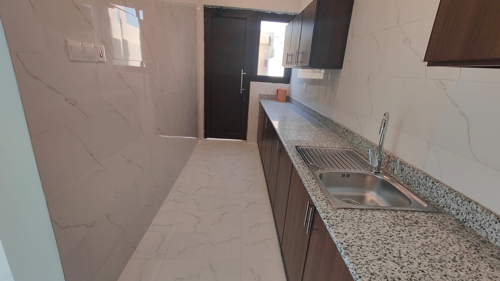 A narrow kitchen with dark wooden cabinets, a granite countertop, and a stainless steel sink. The floor and walls have light-colored tiles. A door with a window is at the end of the room.