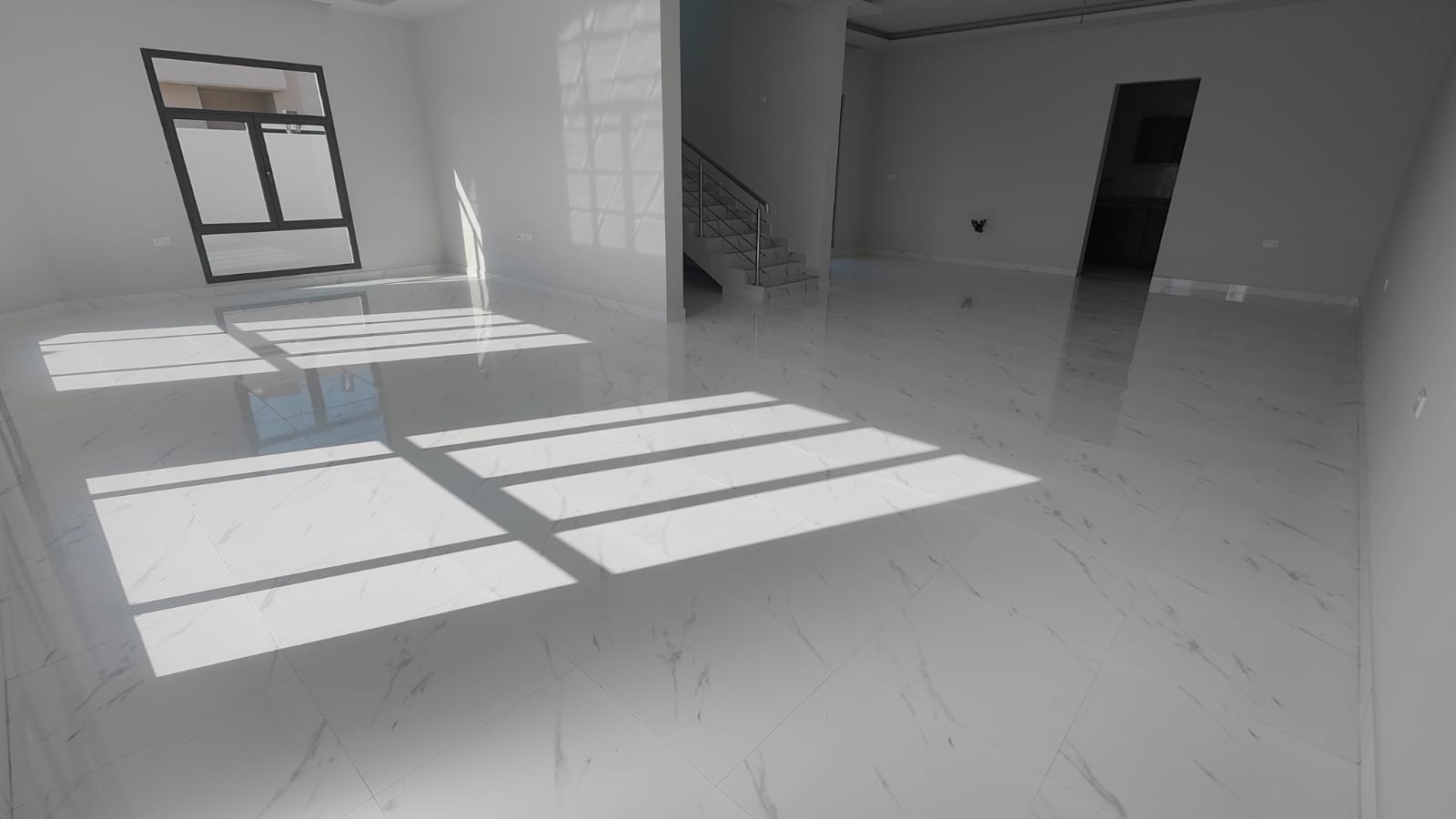 A spacious, bright room with white marble floors, a staircase, large windows casting shadows, and a doorway to another room.