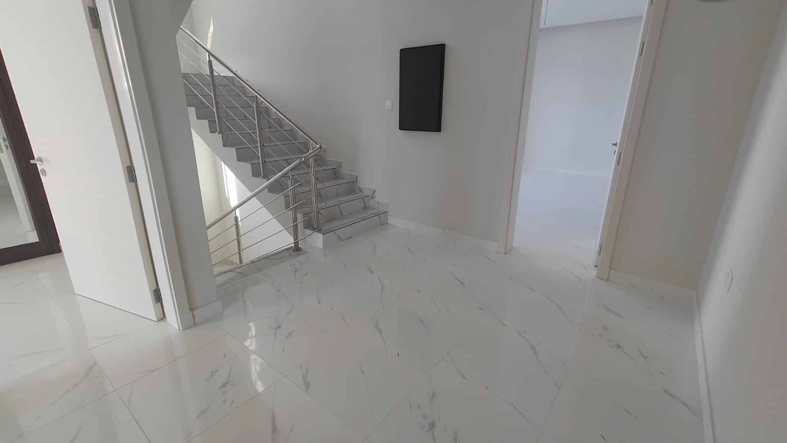 Minimalist interior with light marble flooring, metal rail staircase on the left, and a black rectangular wall art. Two open doors lead to separate rooms.