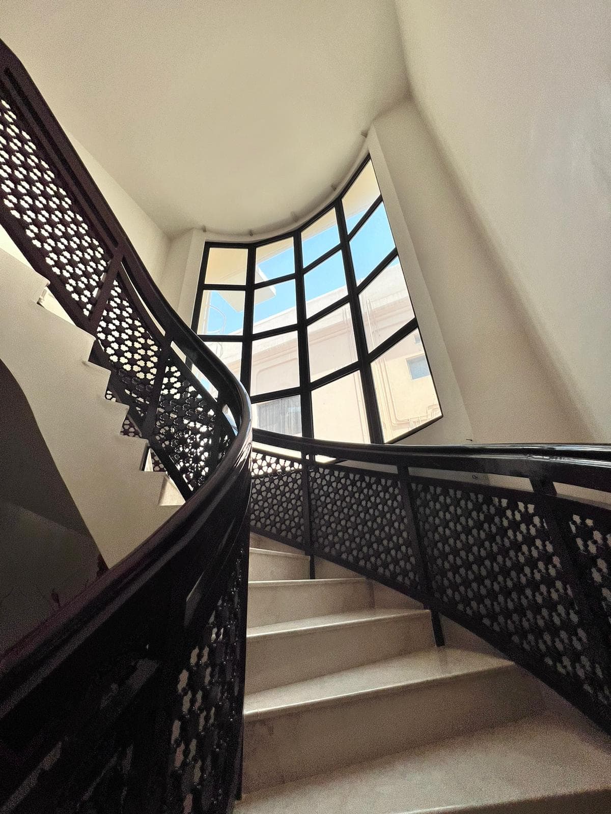 Curved staircase with ornate dark railing, leading upwards to a large, multi-paned window with daylight visible through the glass.