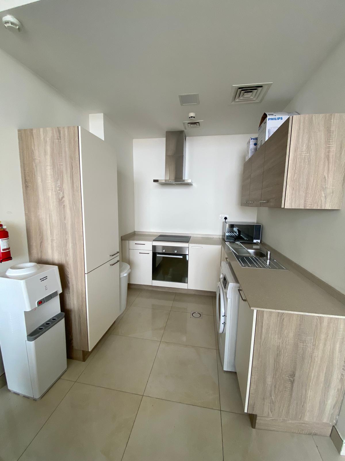 A modern kitchen with wooden cabinets, an oven, a washer, and a stovetop. A water dispenser and a fire extinguisher are on the left. The floor is tiled, and the walls are white.