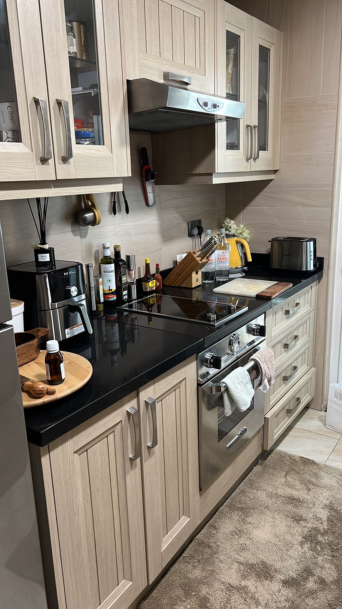 A kitchen with wooden cabinets, black countertop, oven, toaster, and various kitchen appliances and bottles. A towel hangs on the oven handle.