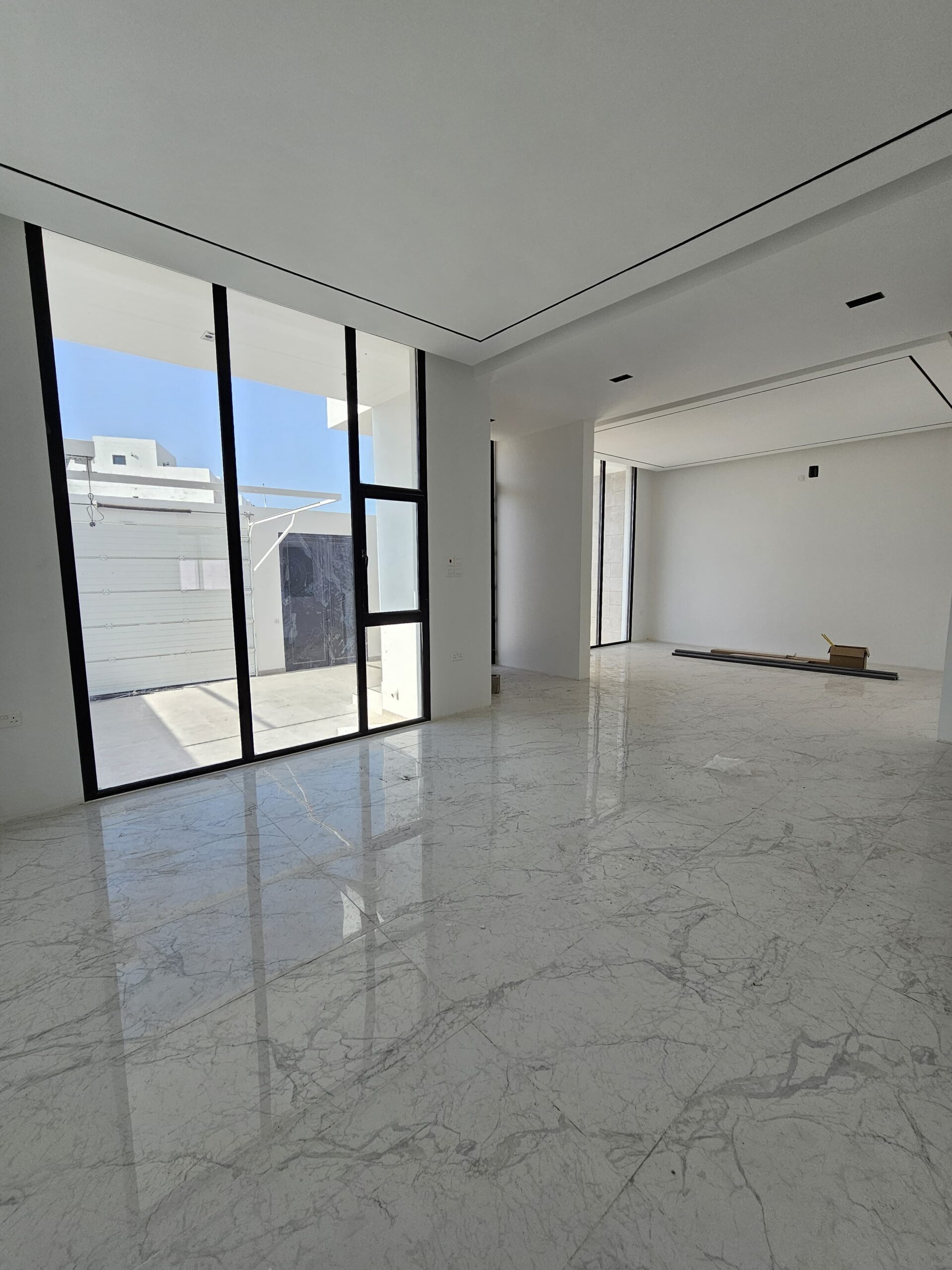 Spacious empty room with large windows, white marble floor, and white walls. A small cardboard box and wooden plank lie on the floor. Bright natural light fills the space.