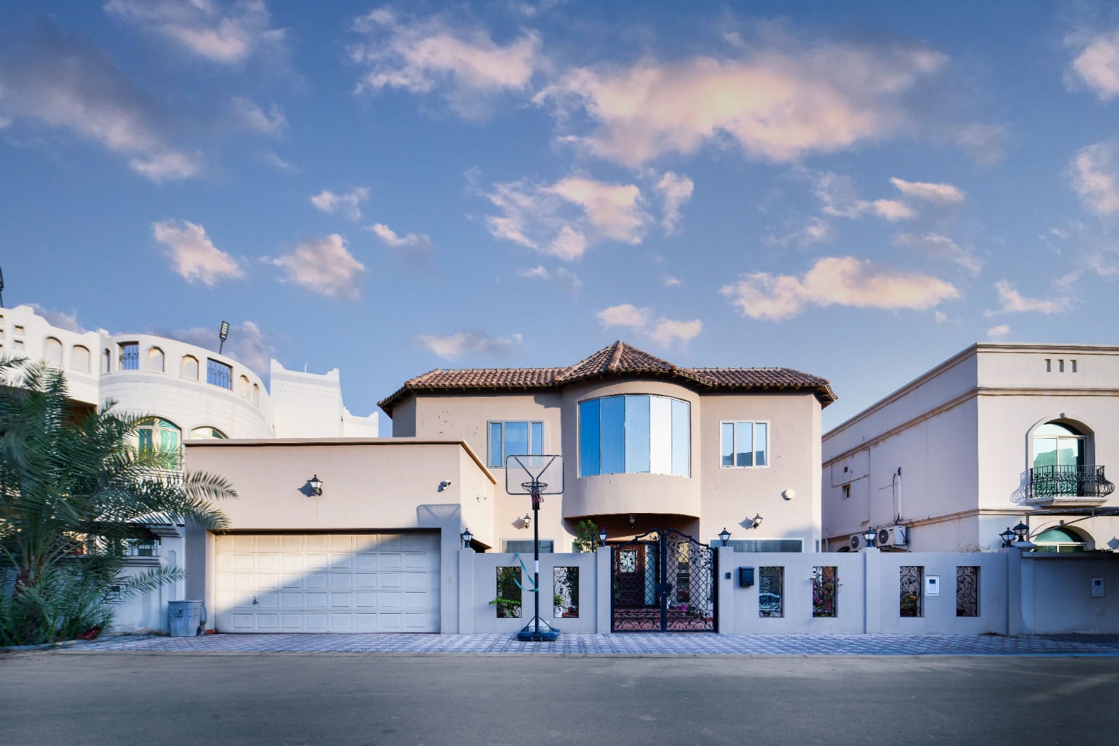 A two-story house with a tiled roof and a basketball hoop in the driveway. It's surrounded by a white fence, with nearby buildings. The sky is partly cloudy.