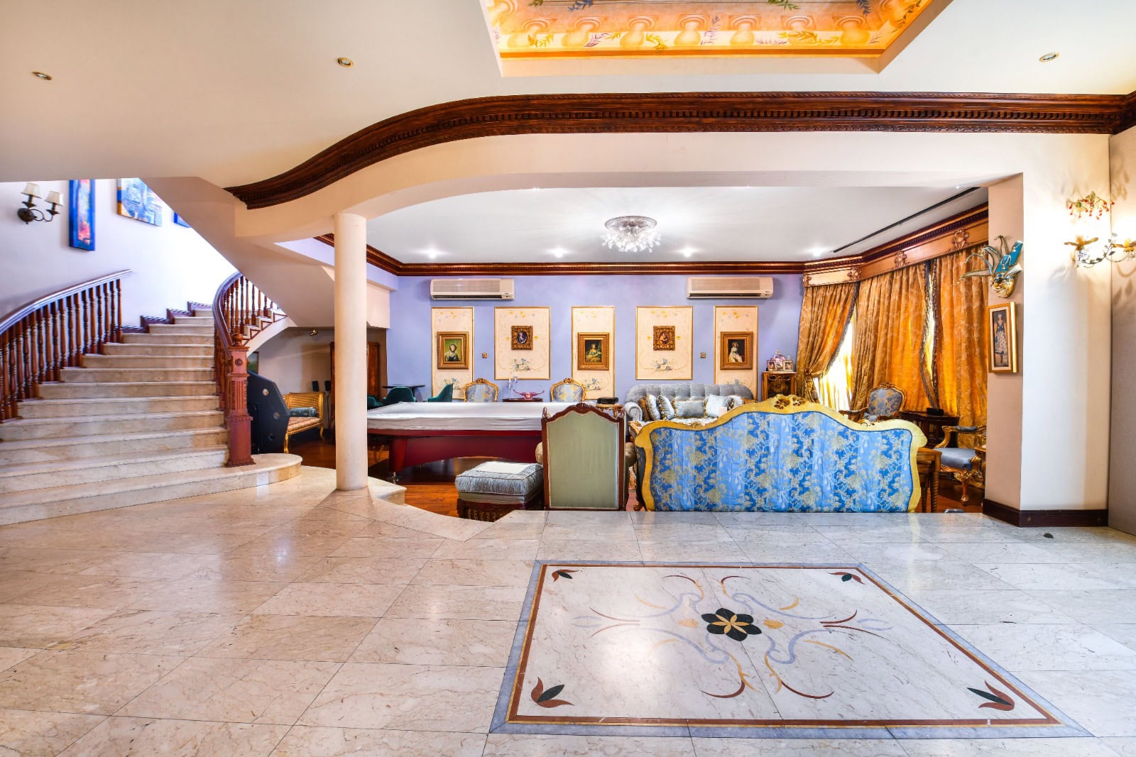 Luxurious living room with ornate furniture, marble flooring, and a grand staircase. Decorative ceiling lights and framed art adorn the walls.