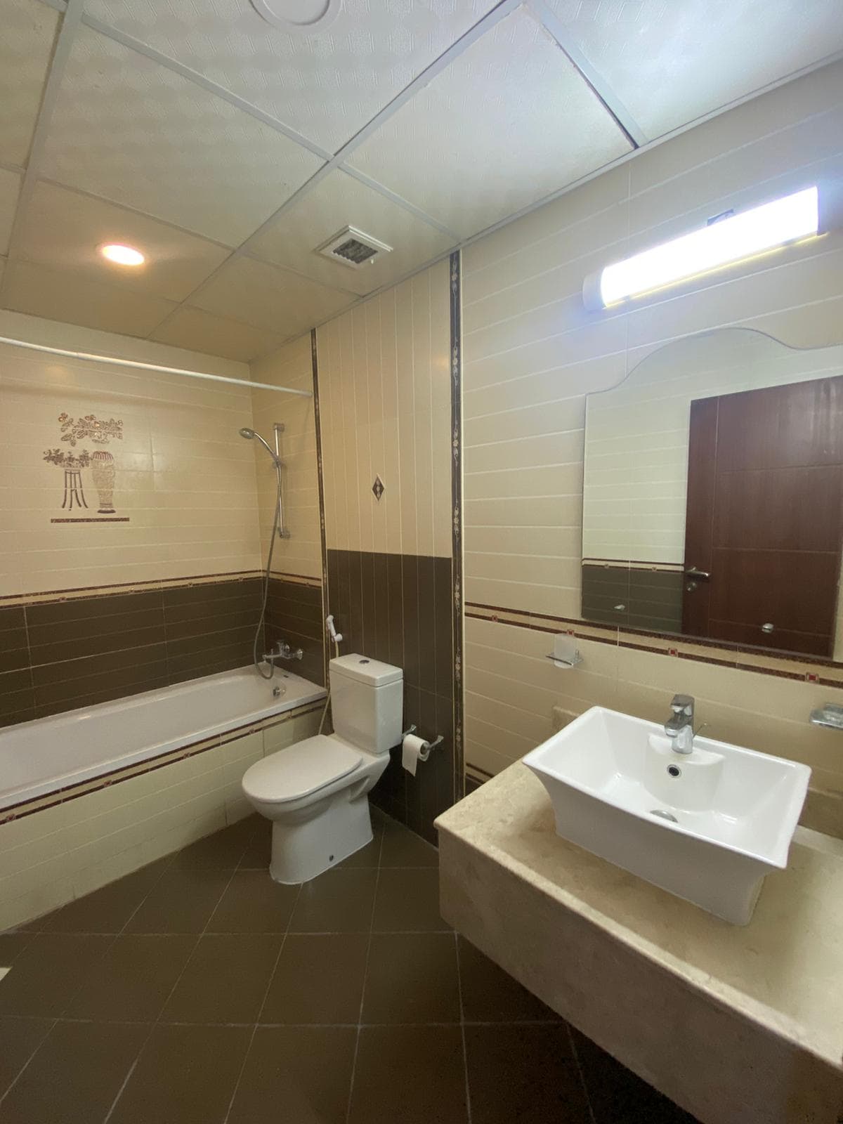 A bathroom with a bathtub, shower, toilet, and sink. There's a wall-mounted mirror above the sink and a light fixture above it. The walls and floor have light and dark tile patterns.