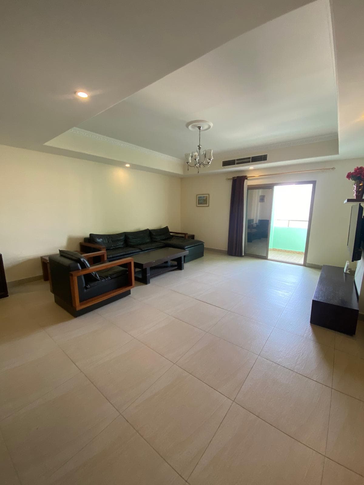 Spacious living room with a black leather sofa set, coffee table, and a TV on a stand. Large window with curtains and a small balcony visible in the background.