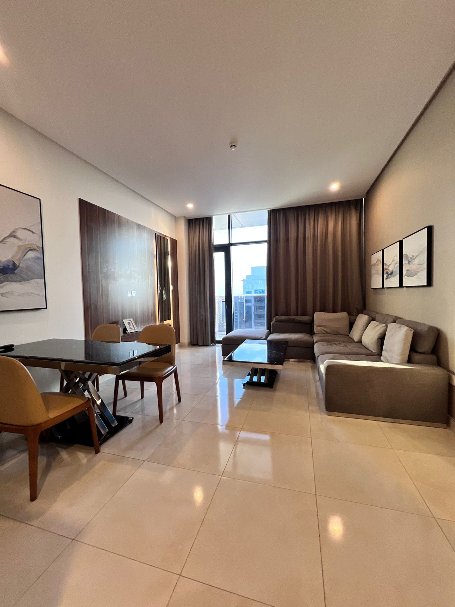 Modern living room with tiled floor, gray sectional sofa, two landscape paintings on the wall, dining area with table and chairs, large window with city view, and brown curtains.