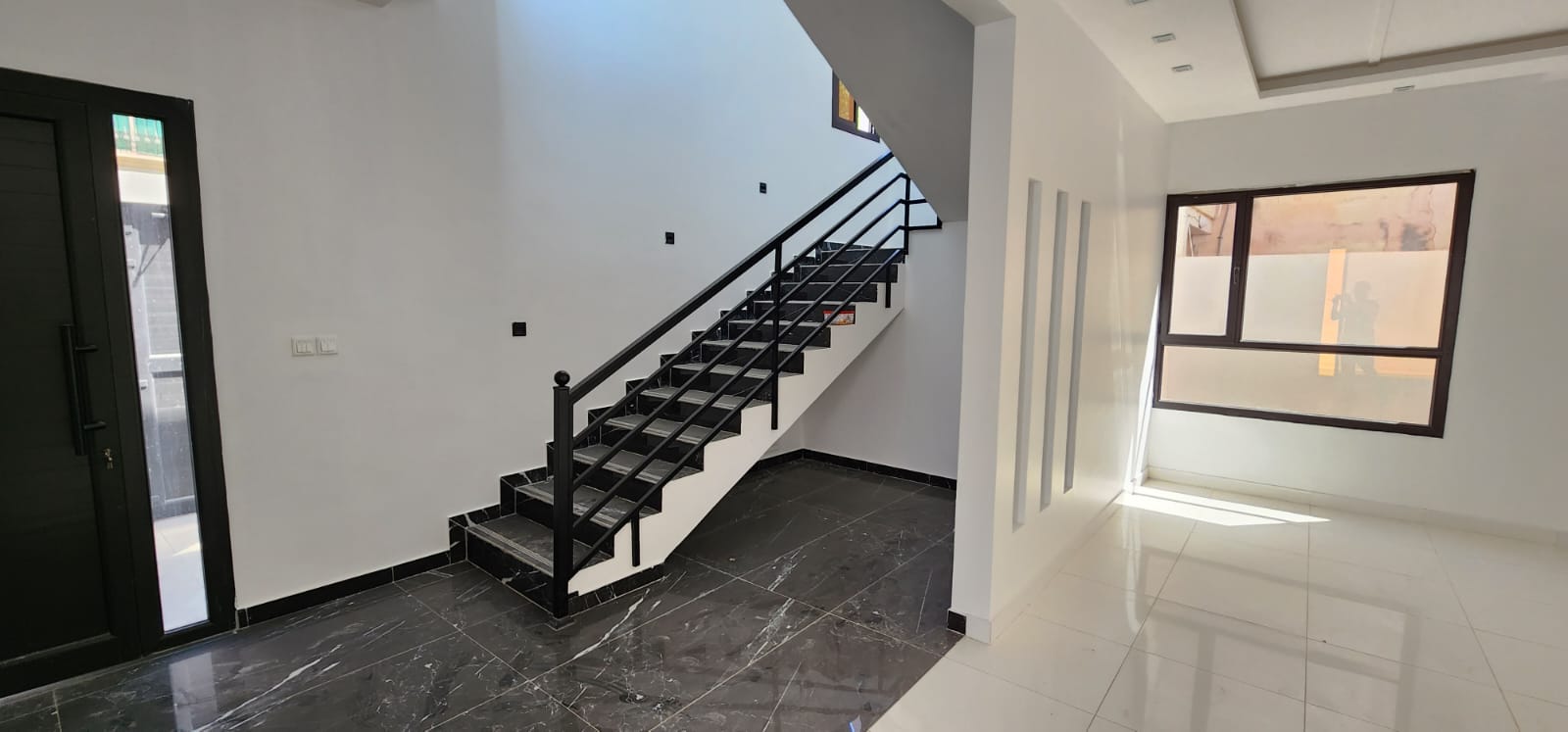 Interior of a modern, empty space featuring a black staircase, black and white tiled flooring, and a large window with natural light.