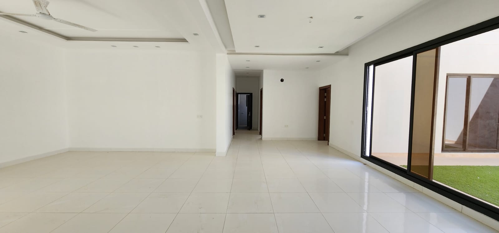 Spacious empty room with white walls, large windows, tiled floor, and a view of a small courtyard.