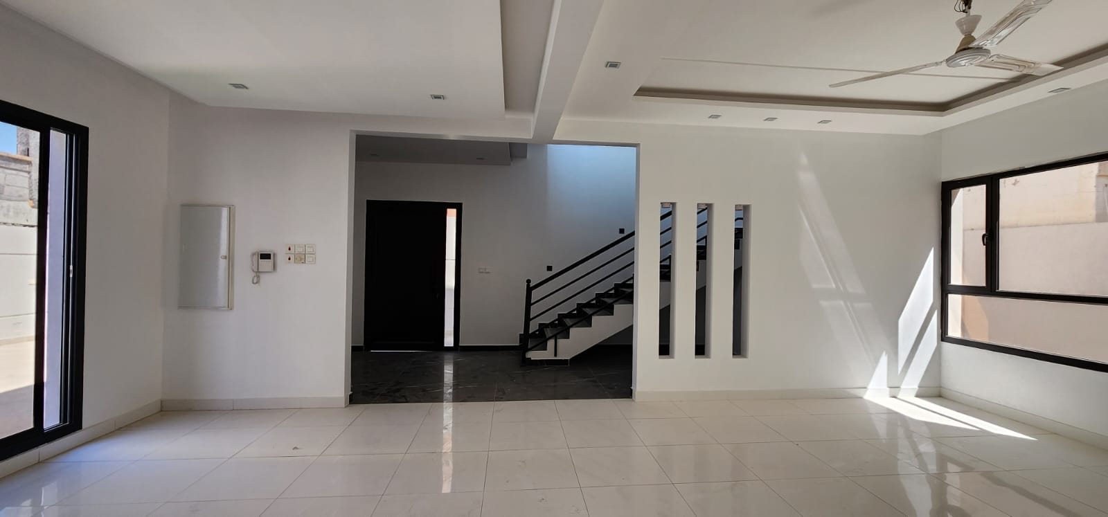 Spacious empty room with white walls, large windows, and tiled floor. A staircase is visible through an open doorway. Ceiling fan and recessed lights are installed.