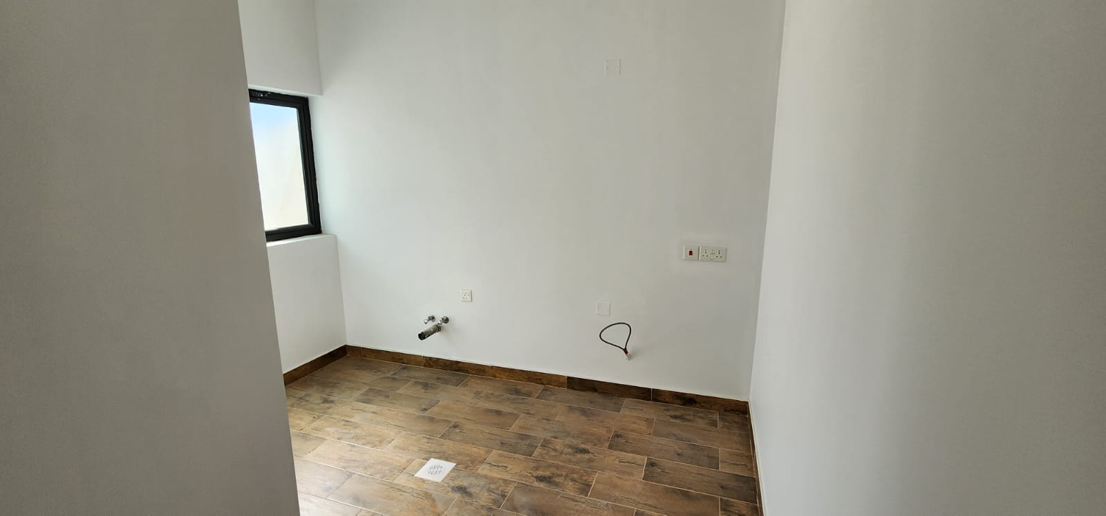 Empty room with white walls, a small window on the left, and wooden flooring. Visible pipes and cables on the walls.