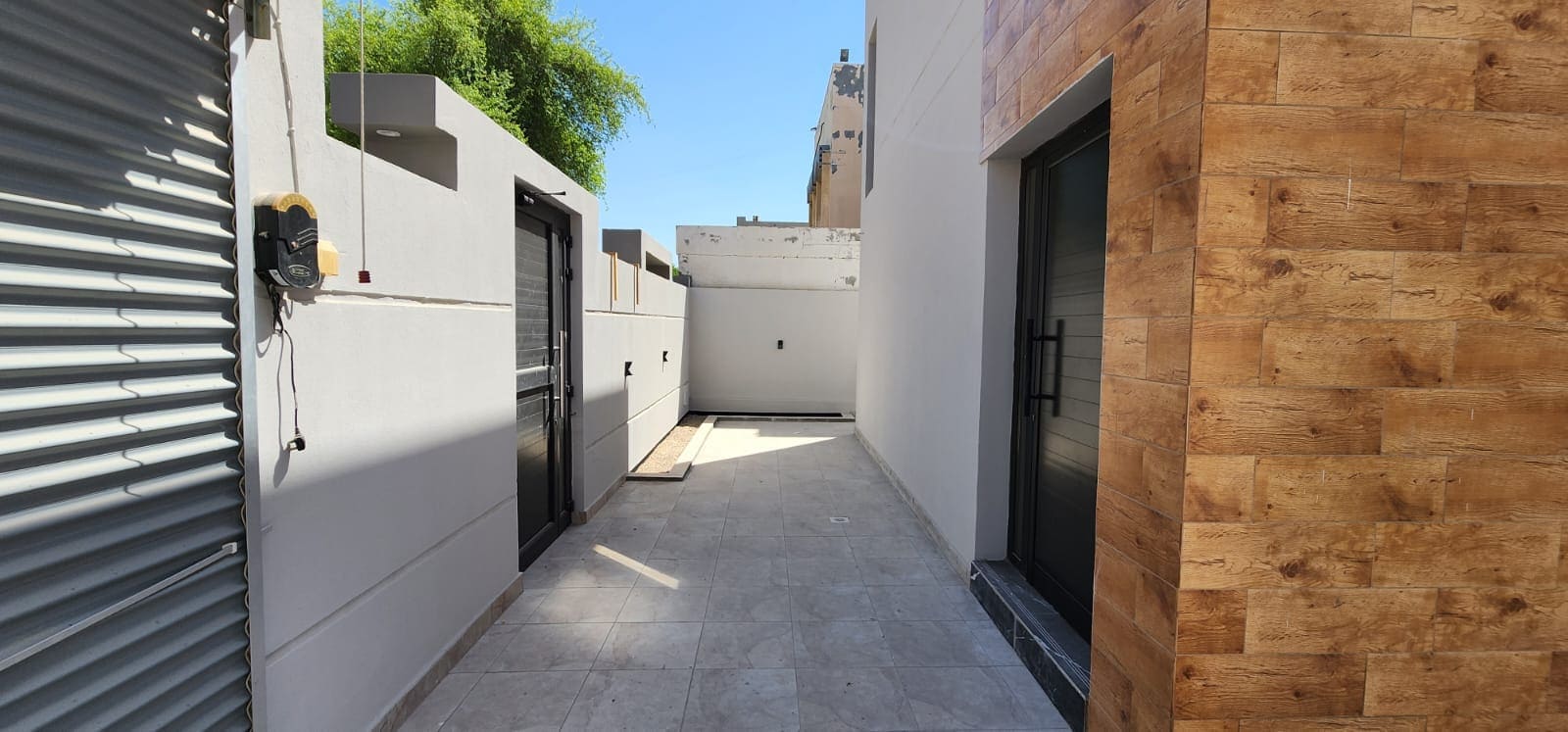 Narrow outdoor passage with tiled floor, flanked by white cement walls, a wooden facade, and a metal shutter door. Bright sunlight illuminates the path.