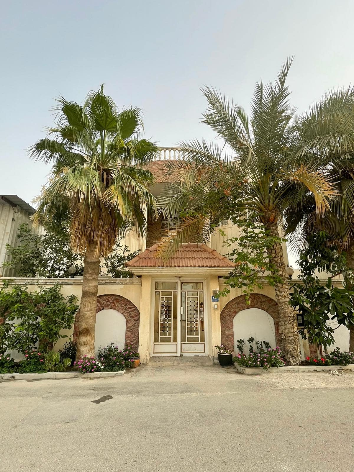 A two-story house with a red-tiled roof, flanked by palm trees, is situated along a street. The entrance features a white door with glass panels and decorative arches.