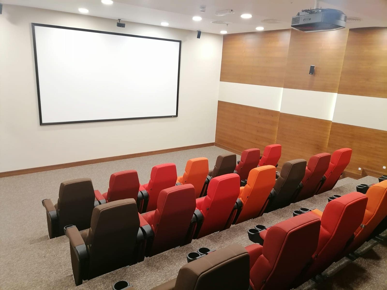 Small theater room with tiered seating in various colors facing a large white screen on a wooden-paneled wall. Ceiling lights are on, and a projector is visible above the seats.