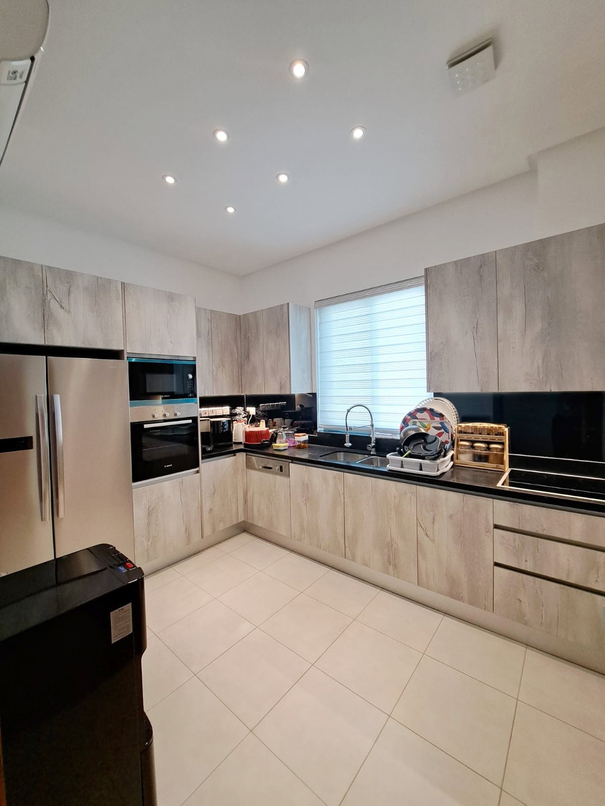 Modern kitchen with wood-pattern cabinets, black countertops, and white tile floor. Includes built-in oven, stovetop, sink, and a window with a white blind.