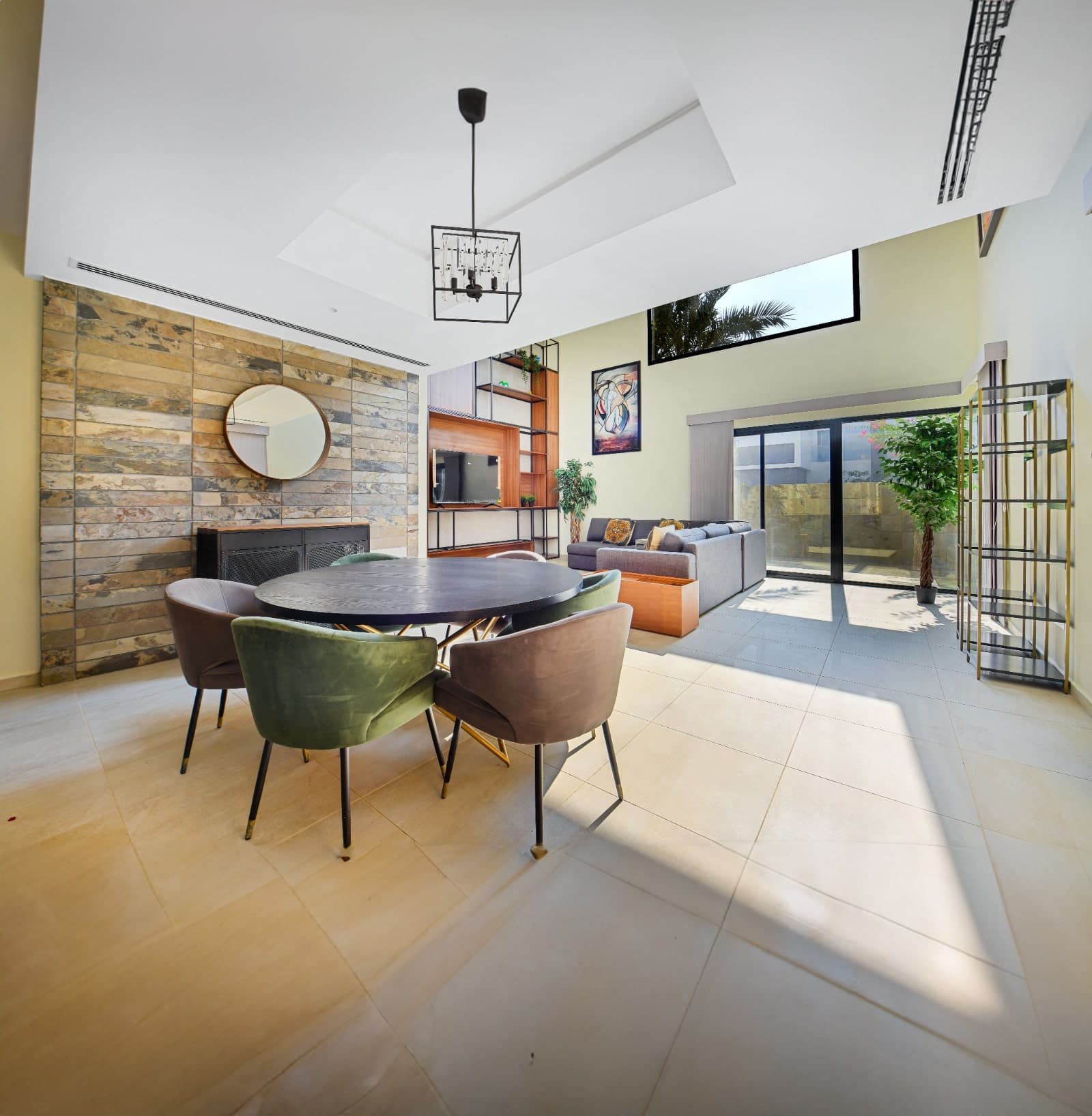 Modern open-plan living area with round dining table, chairs, and a lounge. Stone feature wall, large windows, and bright natural light.