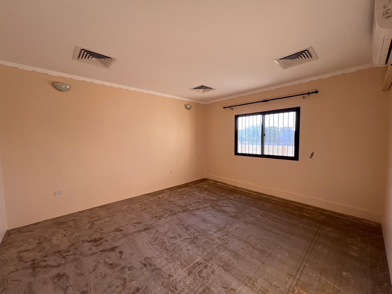 Empty room with beige walls and carpet, a window with bars, two wall-mounted lights, air conditioning unit, and two ceiling vents.