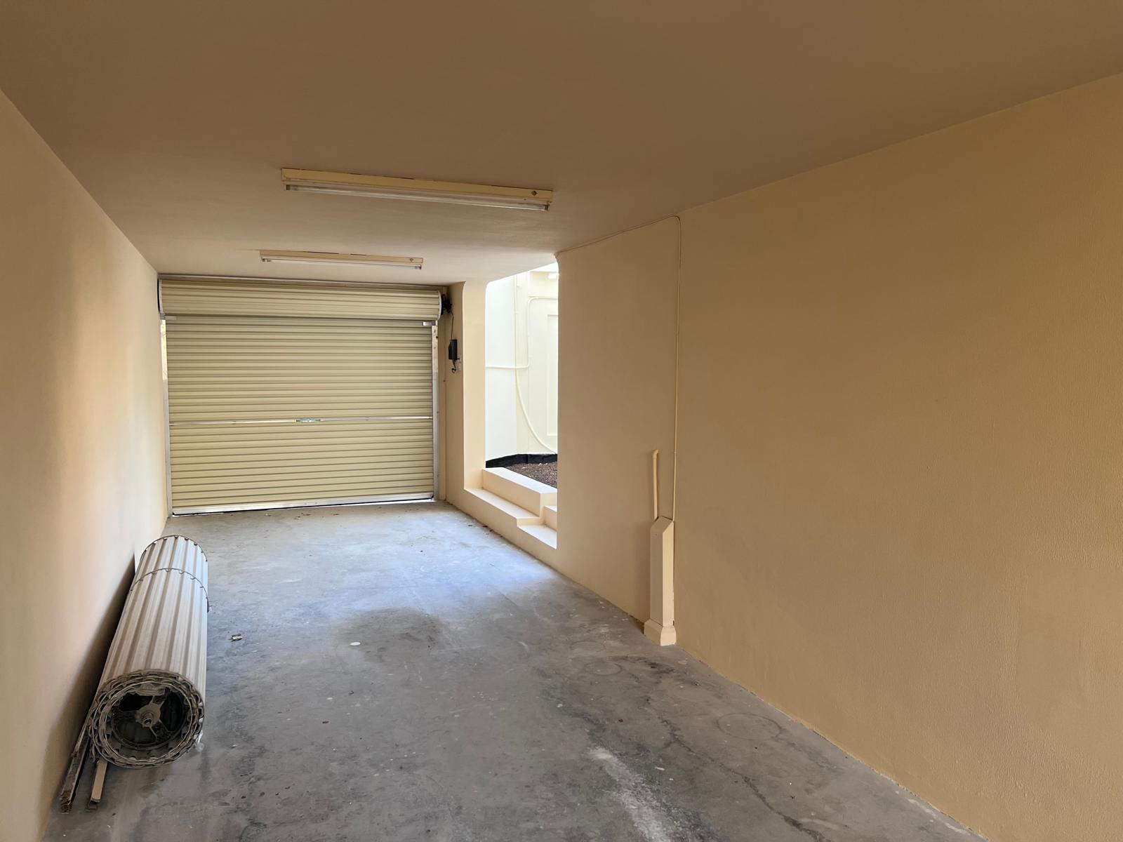 Empty garage with a closed roller door, beige walls, and a concrete floor. A rolled-up mat is leaning against the left wall.