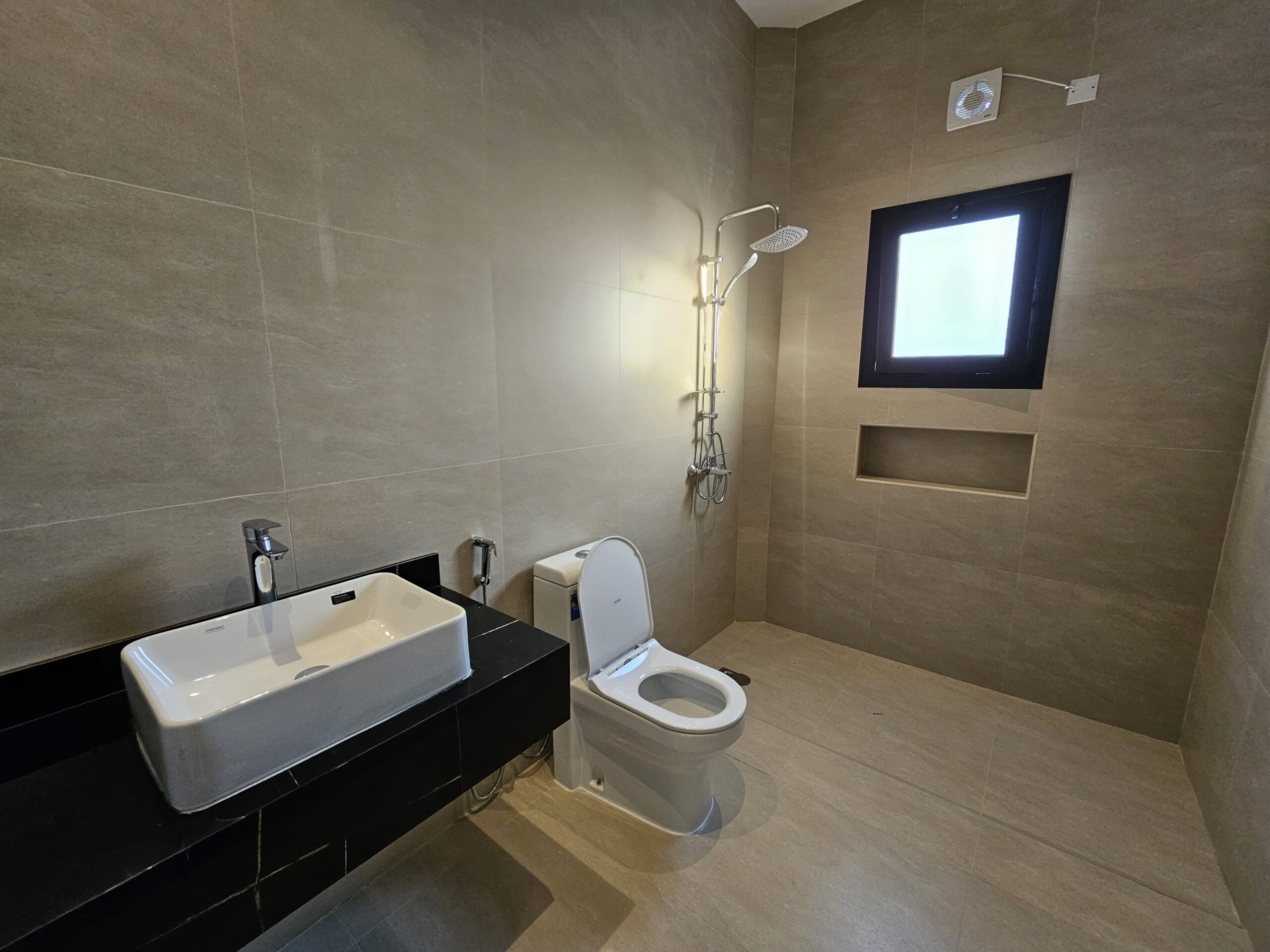 Minimalist bathroom with beige tiles, featuring a white toilet, sink with faucet, wall-mounted shower, and a small window.