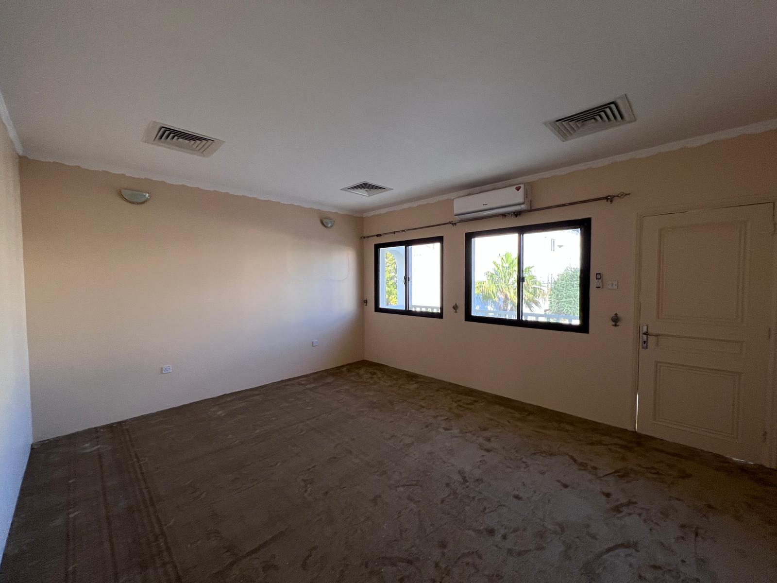 Empty room with beige walls, three windows, a closed door, and ceiling lights. The floor is bare, and an air conditioning unit is mounted above the windows.