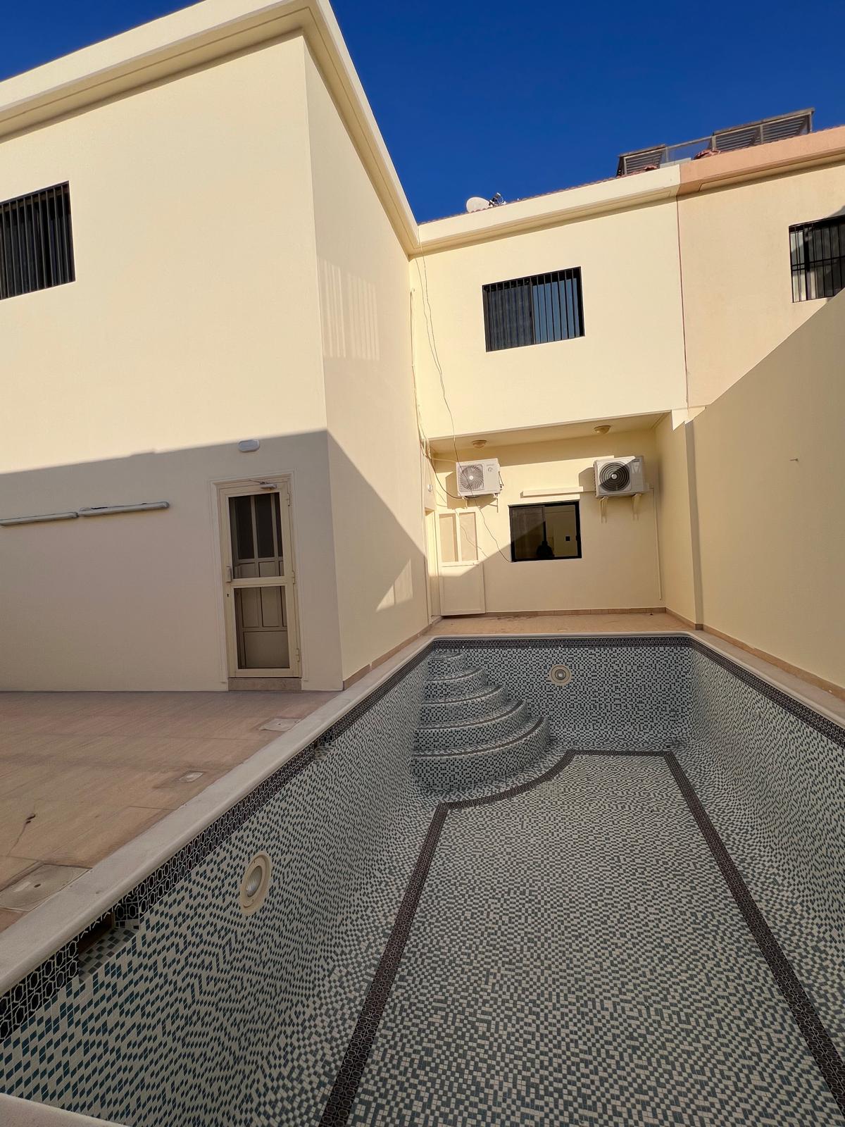 Empty tiled swimming pool in a small courtyard with beige walls and a two-story building. Wall-mounted air conditioners and a clear blue sky are visible.