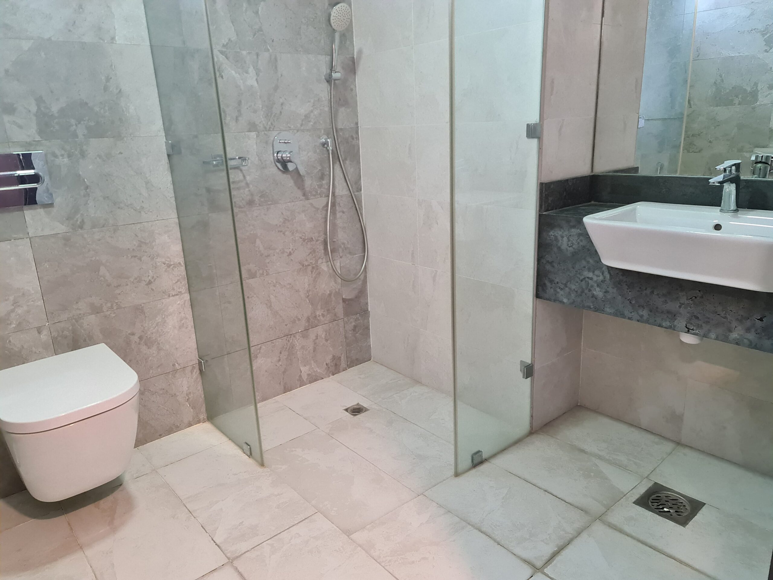 Modern bathroom with a walk-in shower, glass partition, wall-mounted toilet, and sink. Beige tile flooring and walls, with a large mirror above the sink.