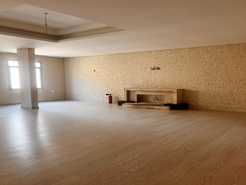 Empty room with light wood flooring, beige textured wall, and two tall windows. A small wooden shelf is mounted on the wall, and a fire extinguisher is placed nearby.