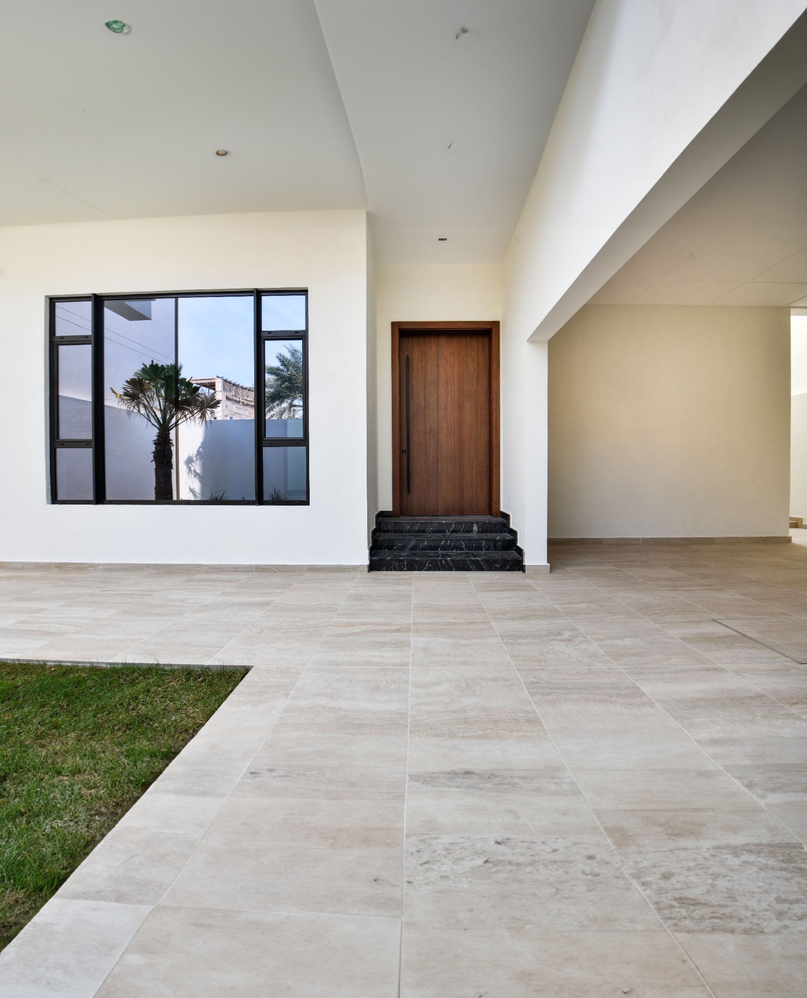 Modern entryway with a wooden door, large window, and tiled floor. Small patch of grass to the left. White walls and ceiling create a clean, minimalist look.