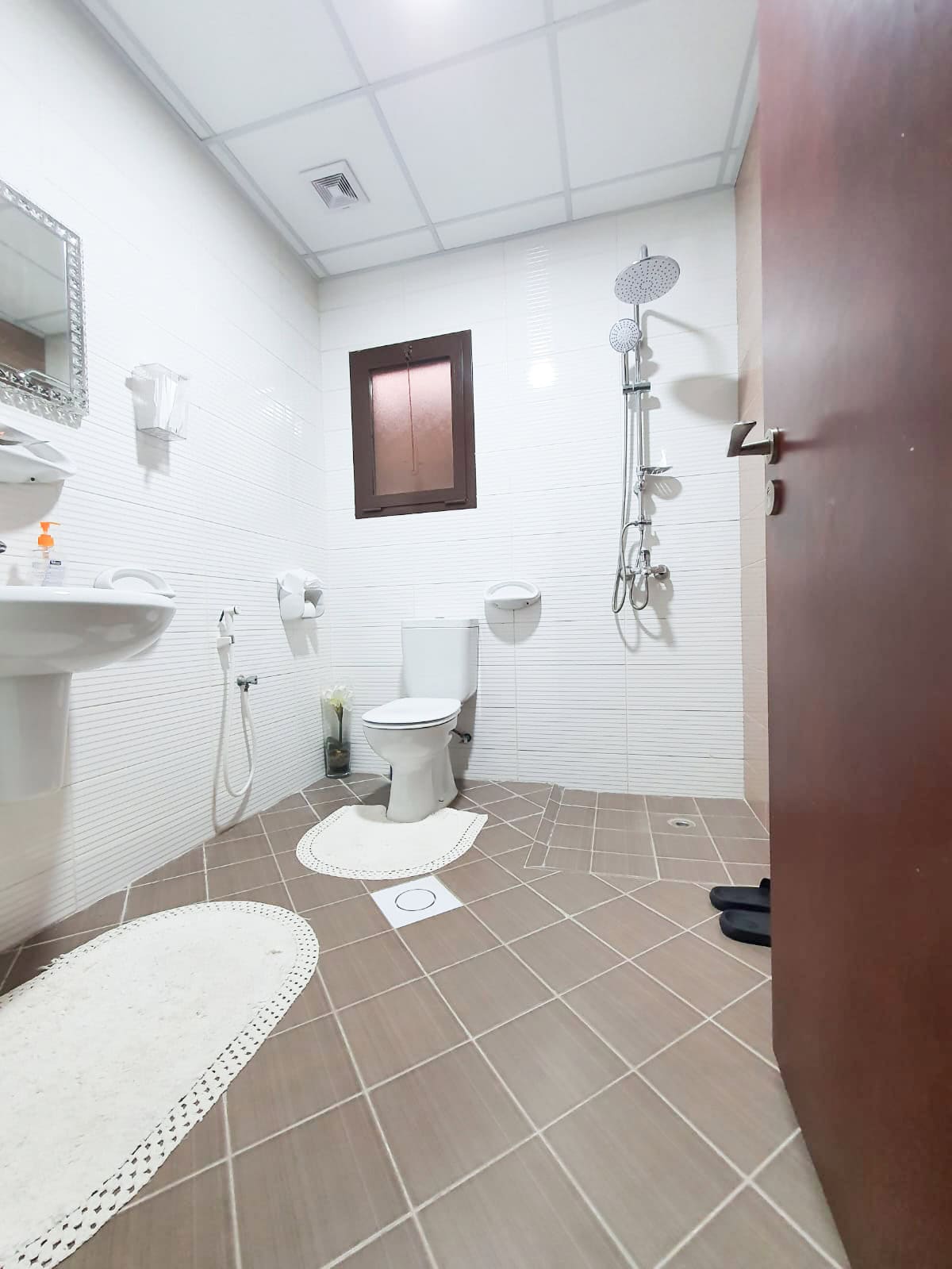 White-tiled bathroom with a toilet, sink, and shower. Brown non-slip mats on tiled floor, wooden door slightly open, small window above toilet, and square ceiling lights.