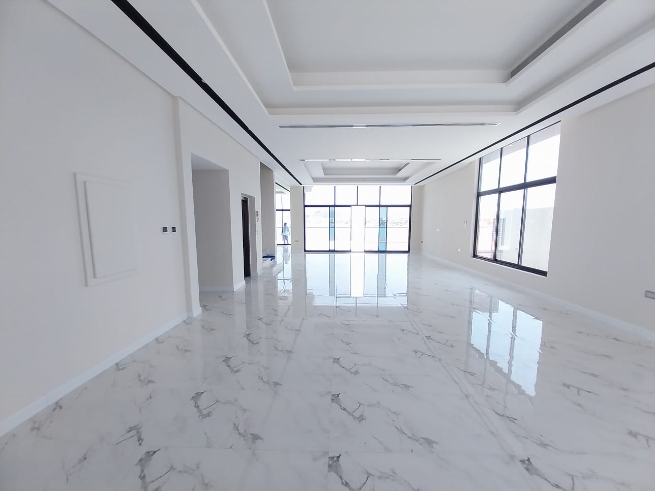 Spacious empty room with marble flooring, large windows, and high ceiling. White walls and ceiling with recessed lighting.