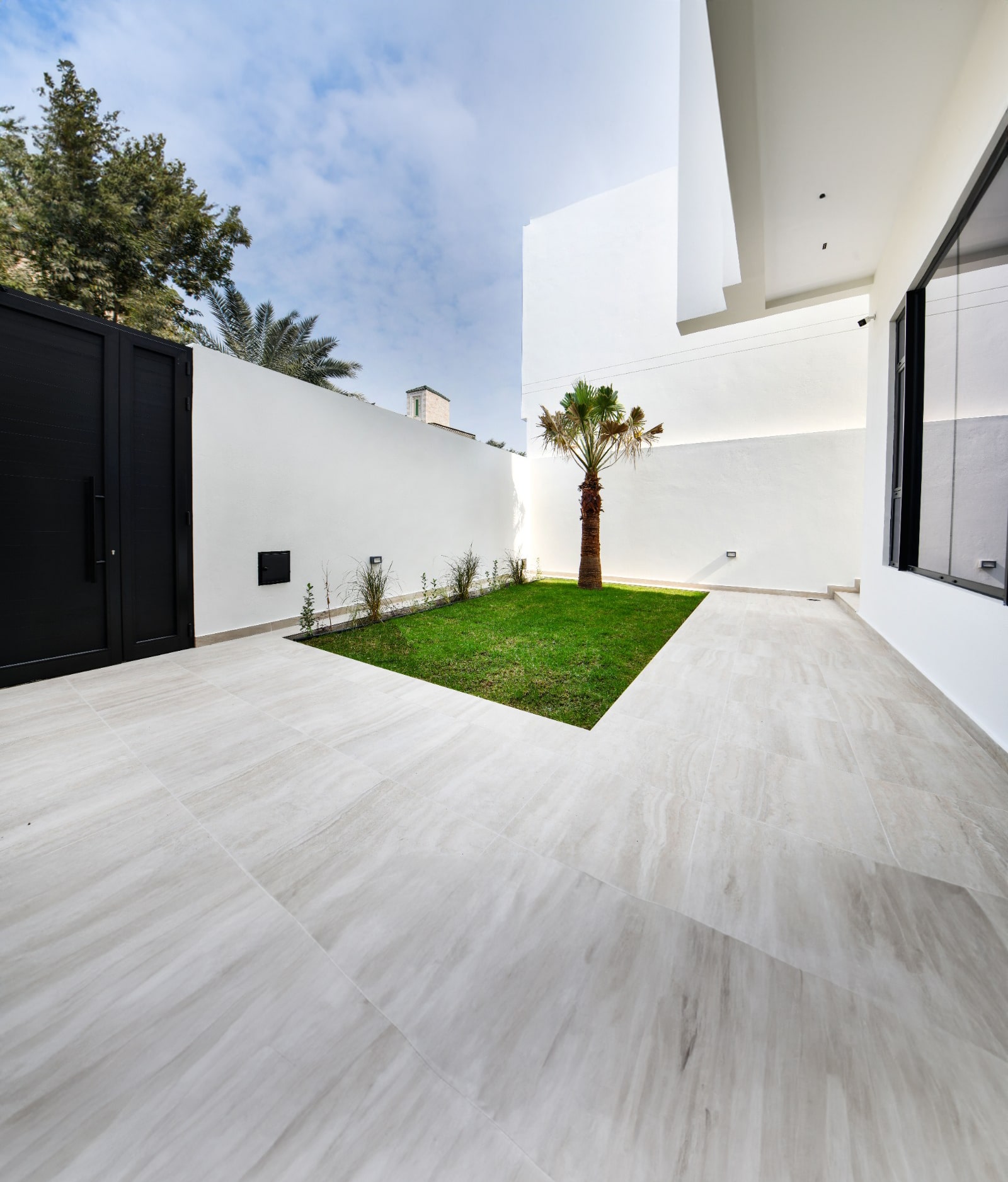 Minimalist courtyard with a small grass patch, a single palm tree, and surrounding white walls. Large beige tiles cover the remaining ground area, with a black gate on the left.