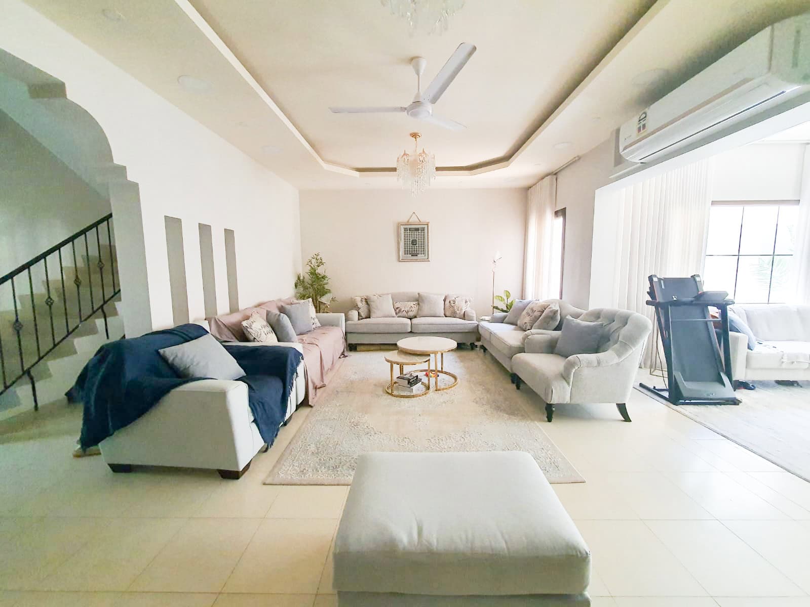 A spacious living room with a U-shaped sectional sofa, a treadmill, and a ceiling fan. A chandelier hangs from the ceiling, and there's a large window with sheer curtains on the right.