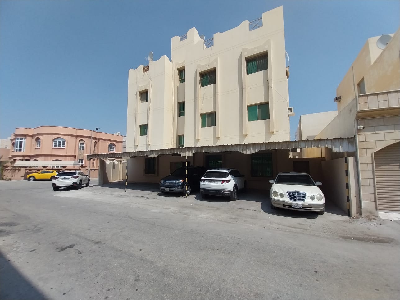 A three-story beige apartment building with green windows. Four cars are parked under an awning in front, and neighboring houses are visible under a clear blue sky.