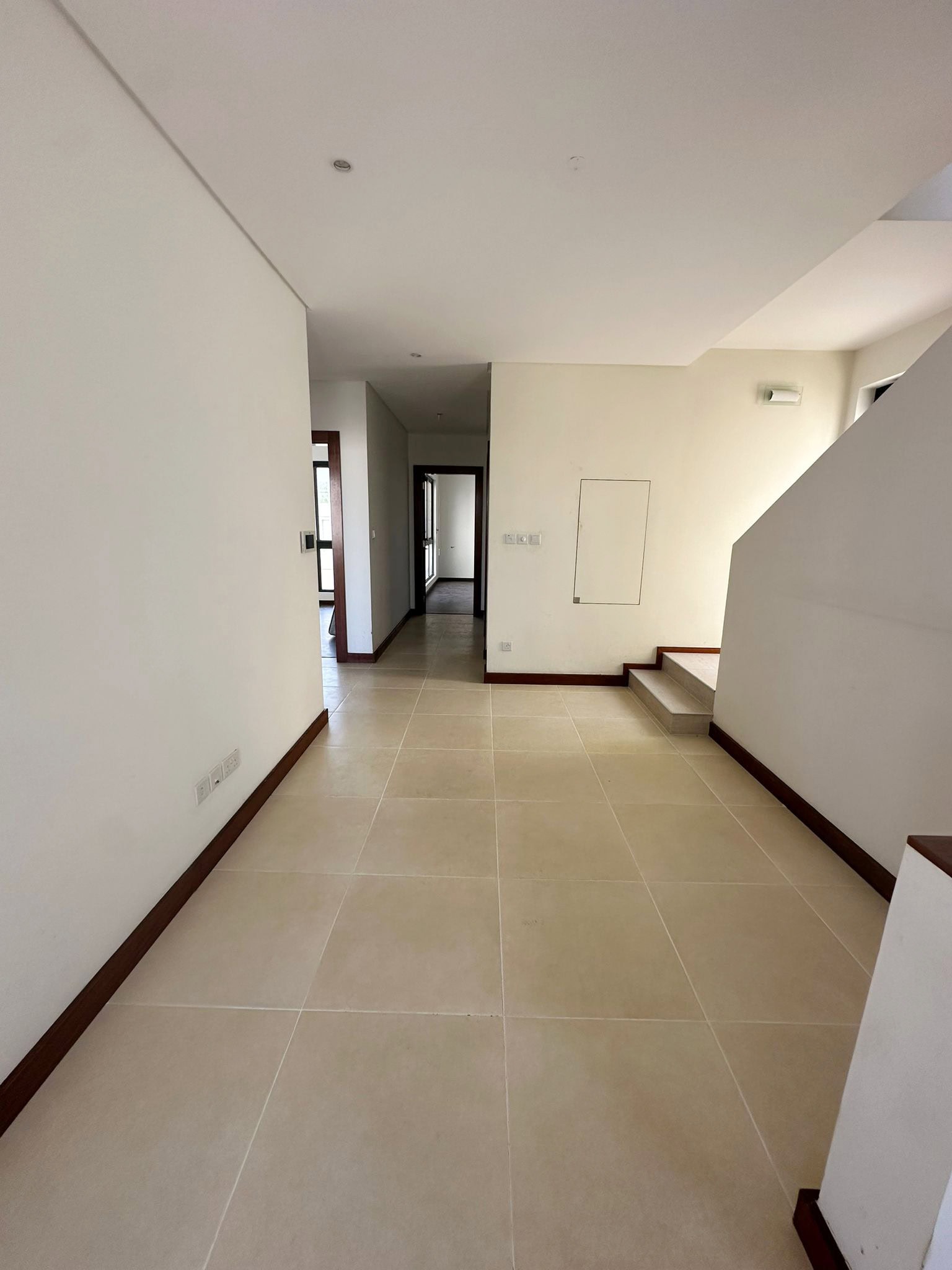 Spacious room with light beige tiled floor, white walls, a staircase, and doorways leading to other areas.