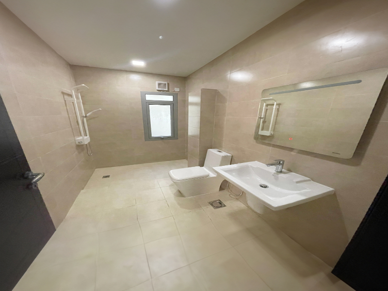 Spacious bathroom with beige tiles, a wall-mounted sink, a toilet, and a shower area. A large mirror is above the sink. There is a window on the far wall.