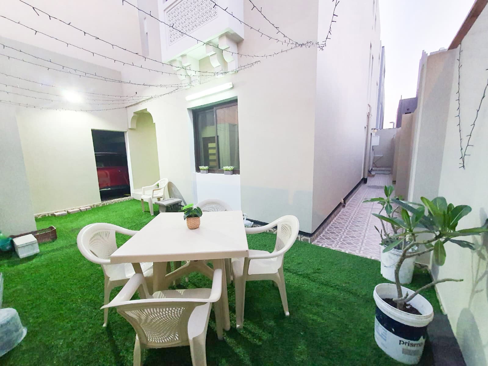 A small outdoor patio with a white table and four chairs on artificial grass, surrounded by beige walls. A potted plant is on the table and string lights hang overhead.