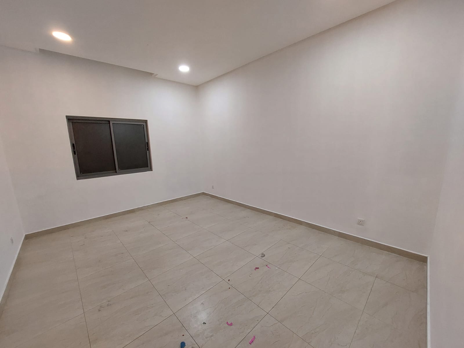 Empty room with tiled floor, white walls, a single window with black frame, and ceiling lights.