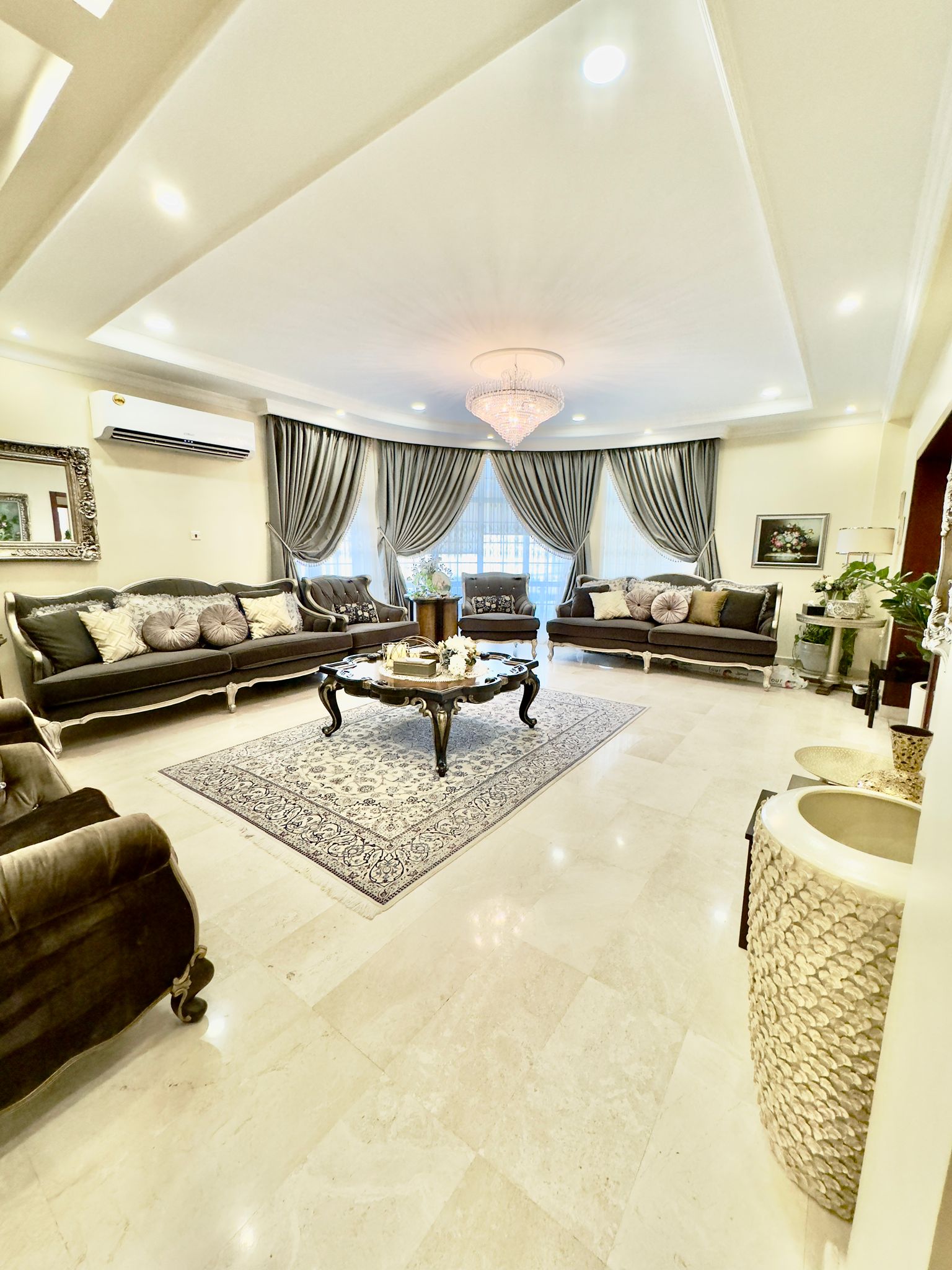 Elegant living room with two dark leather sofas, a central coffee table on a patterned rug, a chandelier, and large windows with curtains. Indoor plants and wall art enhance the decor.