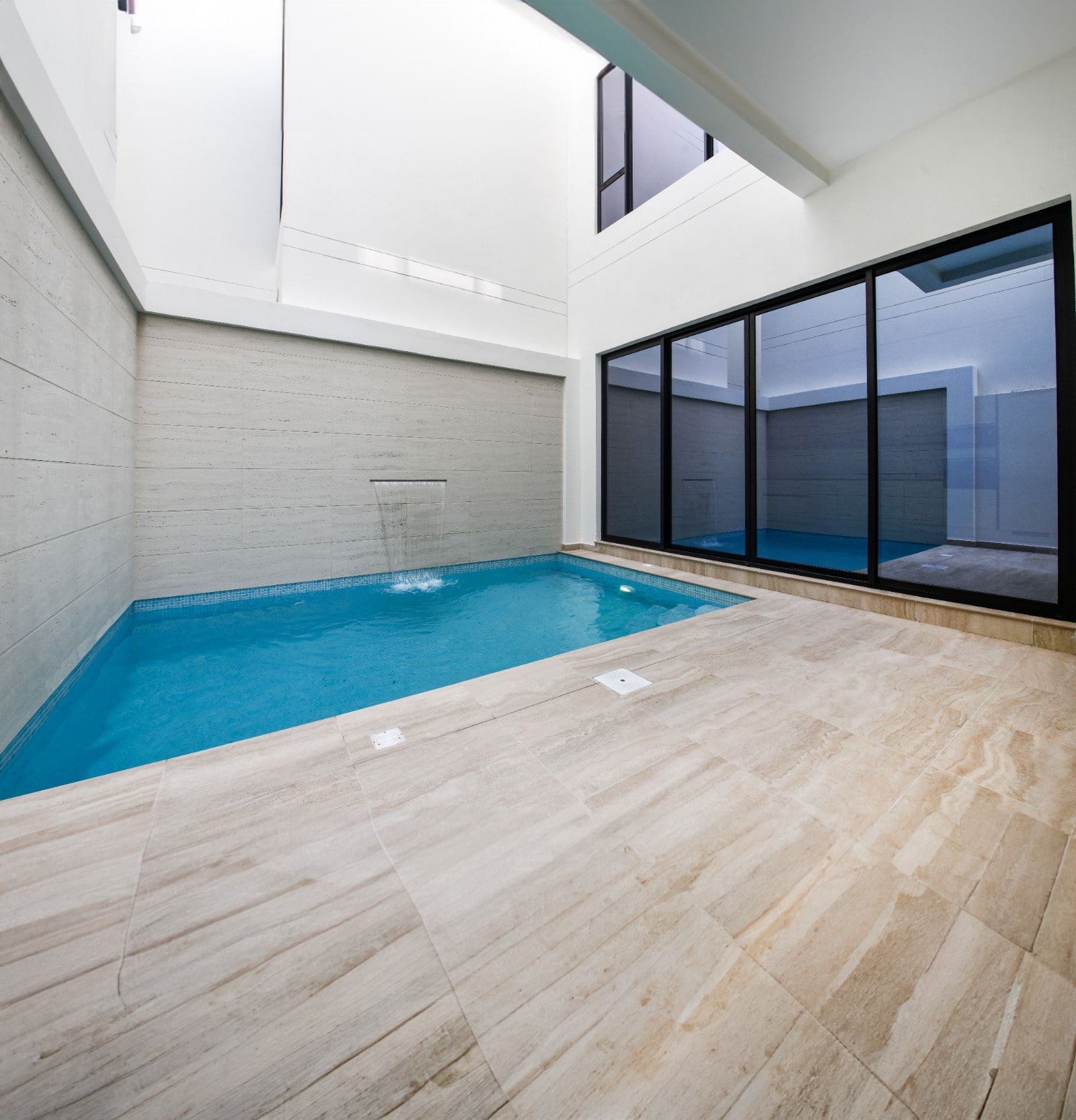 Indoor swimming pool with a waterfall feature, surrounded by beige stone tiles, and large glass windows.