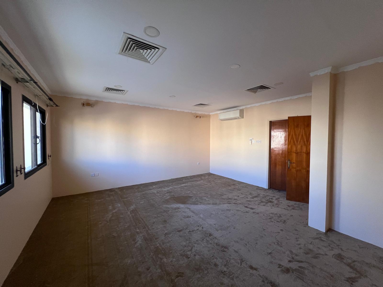 Empty room with beige walls, carpeted floor, ceiling lights, air vents, and air conditioning unit. Windows line one wall and a wooden door is present.