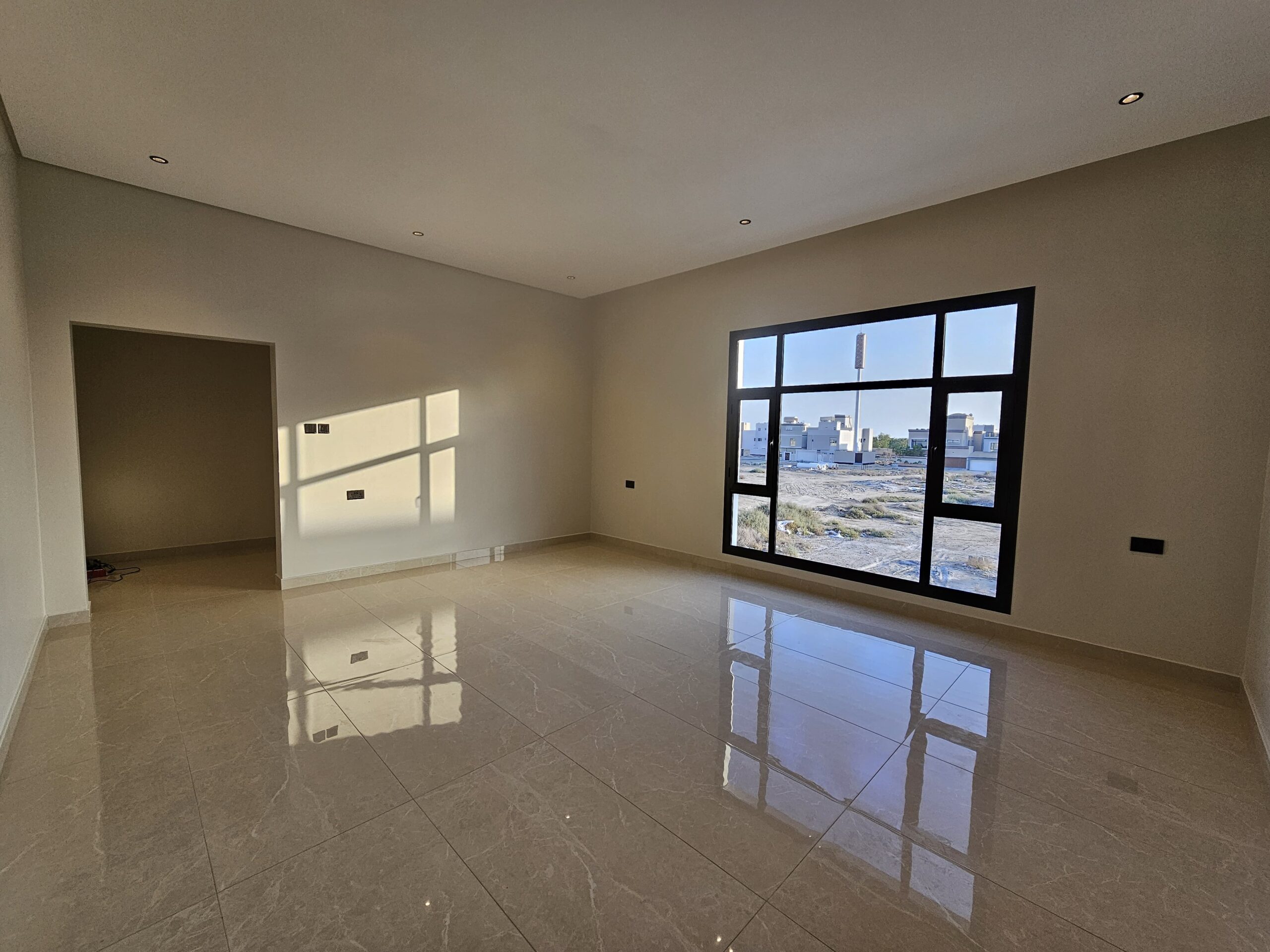 Empty room with beige tiled floor, cream walls, and a large window overlooking a cityscape. Ceiling lights are recessed and natural light enters through the window.