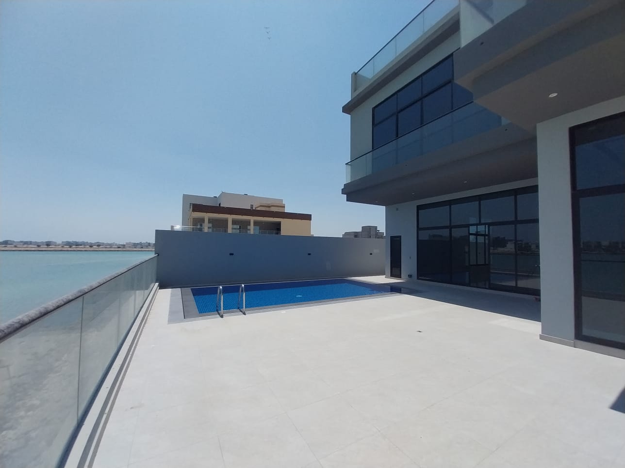 Modern beachfront house with a glass railing, a small pool on the patio, and clear skies. Ocean view on the left side.