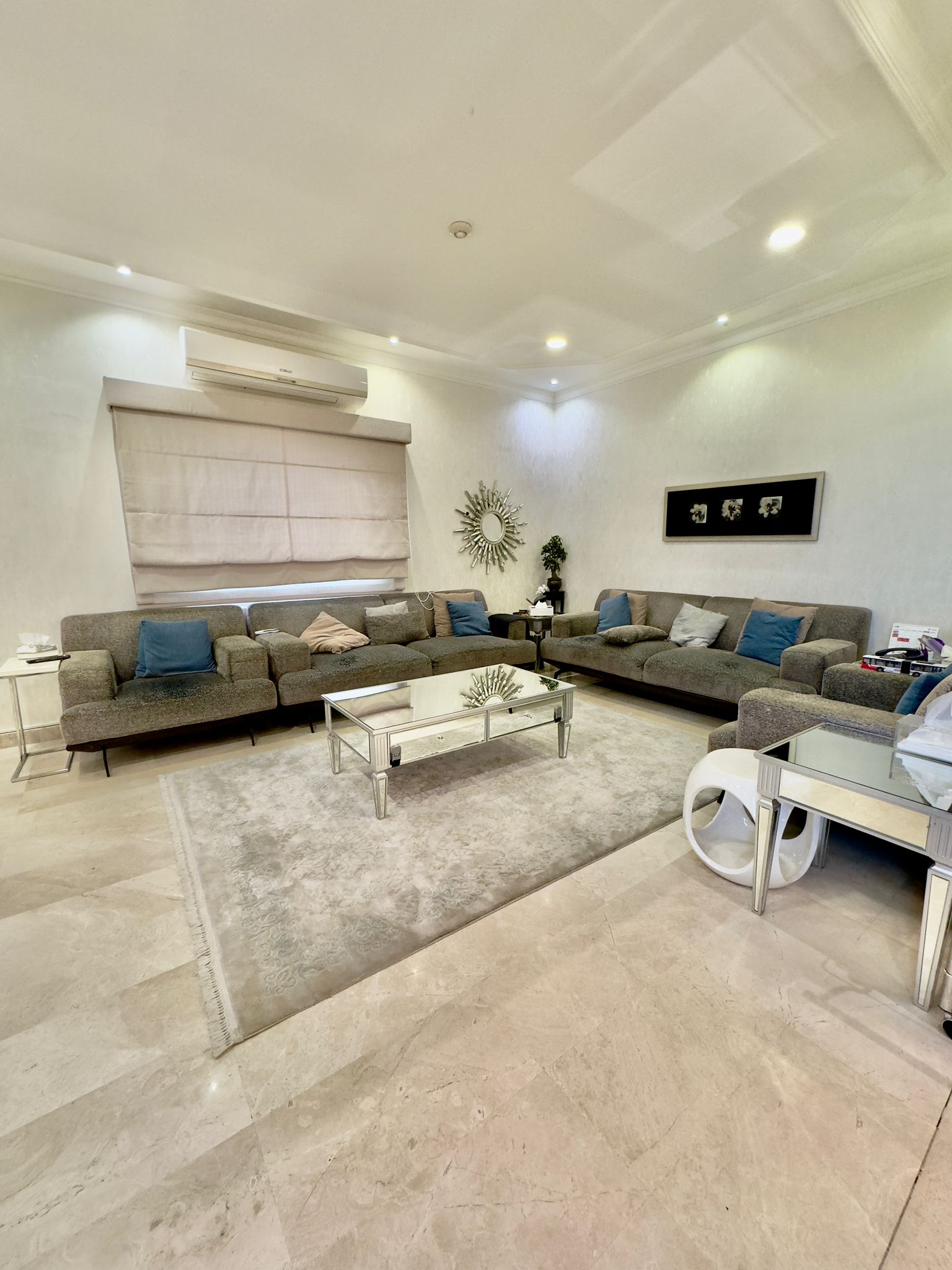 A modern living room with two gray sofas, a glass coffee table, a large rug, and wall decor including a sunburst mirror and framed art. Neutral tones dominate the space.