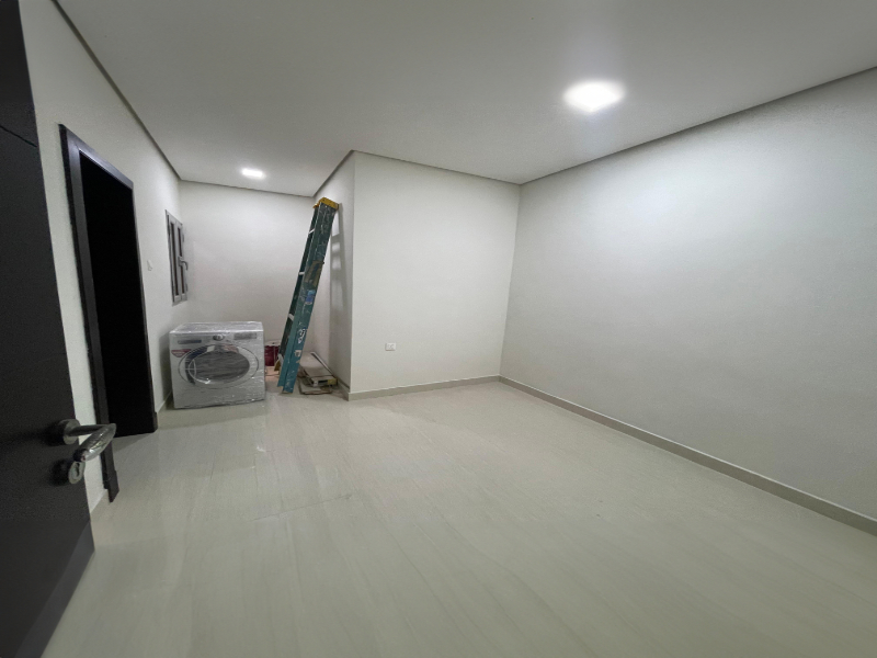 Empty room with white walls and light gray floor. A ladder leans against the back wall next to a boxed washing machine. A black door is partially open on the left.