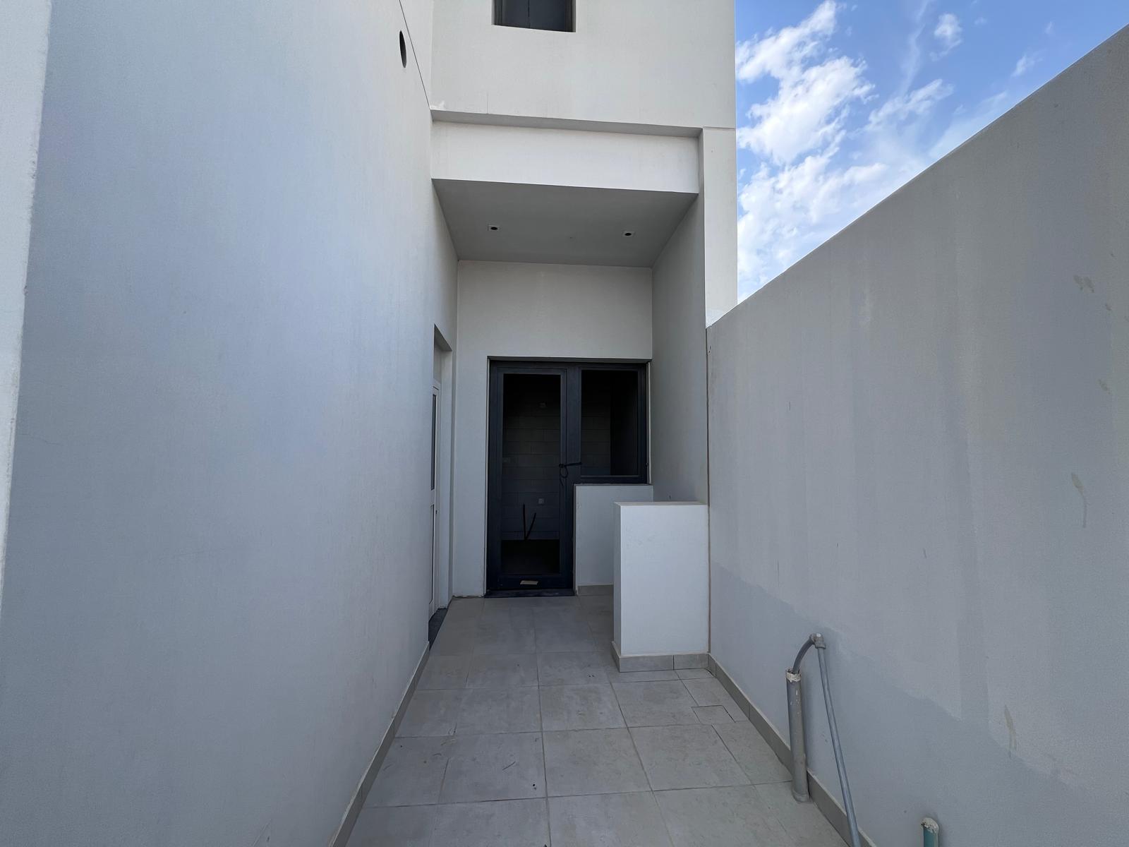 Narrow outdoor corridor with tiled floor, white walls, and an entrance door at the end. Blue sky with clouds visible above.