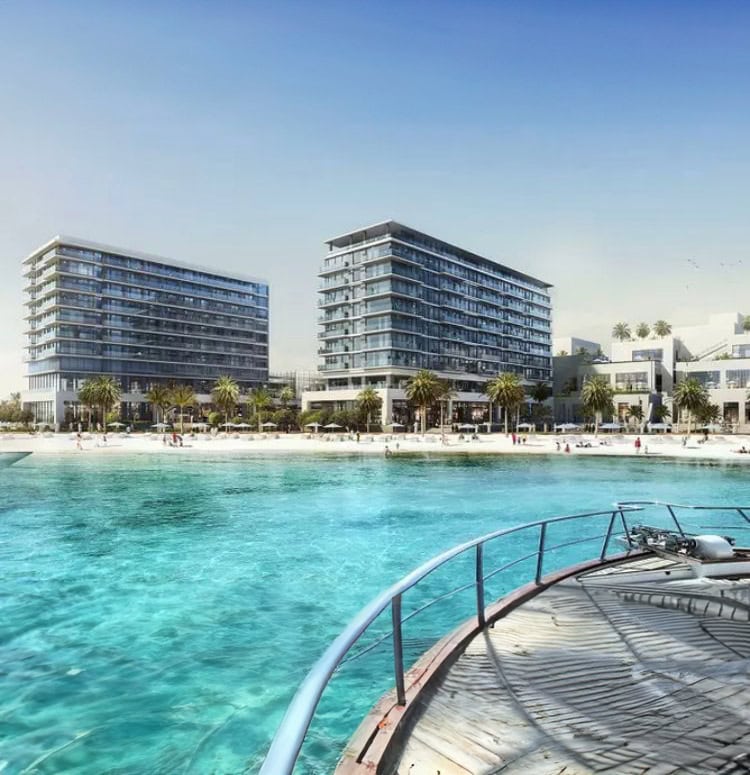 View from a docked boat showing clear blue waterfront, sandy beach, and two modern multi-story buildings surrounded by palm trees under a clear sky.