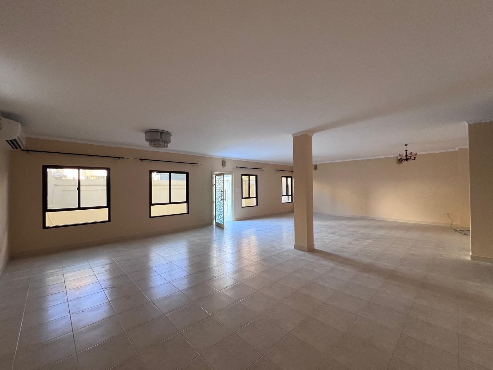 Spacious empty room with tiled floors, beige walls, and multiple windows. A door leading outside is visible. Ceiling lights are installed.